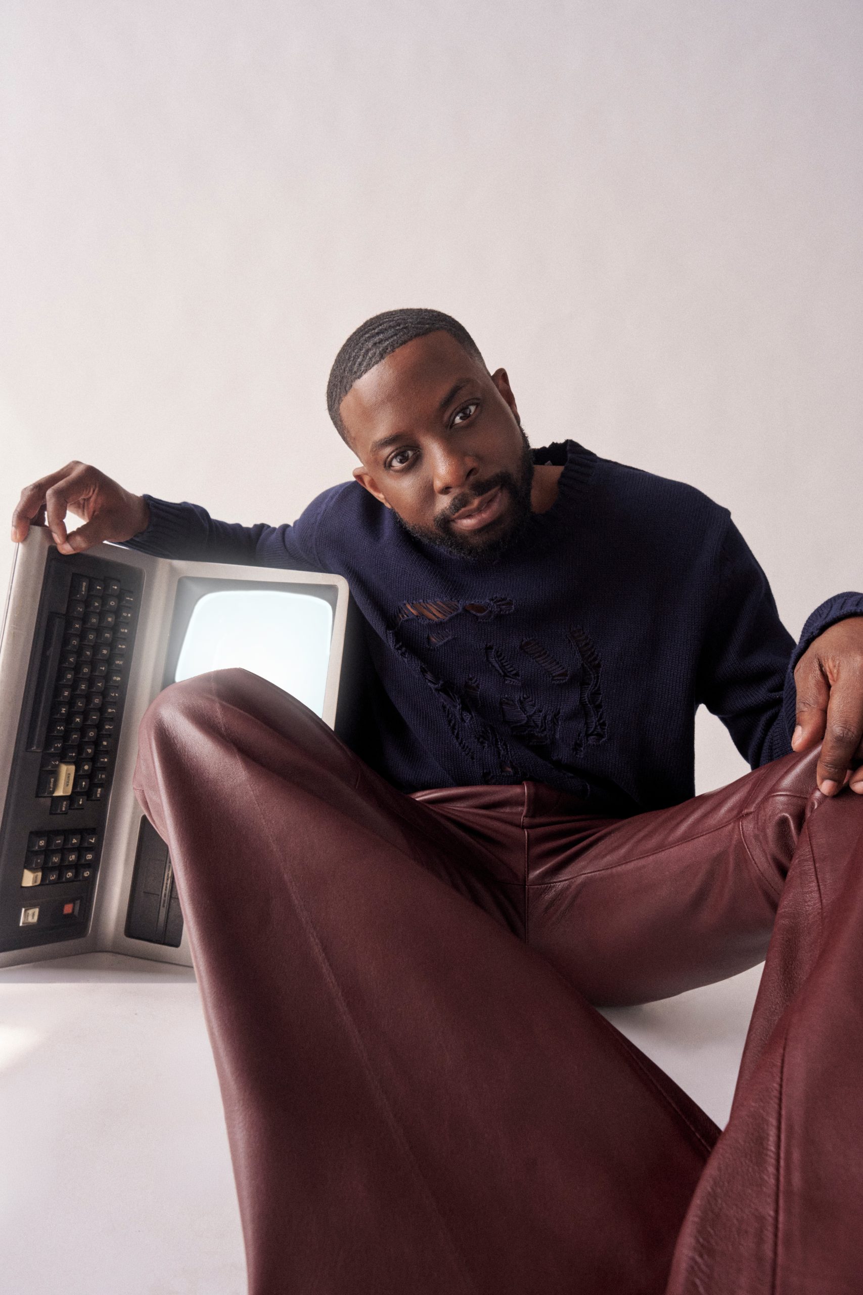 John Imah seated in a leather jacket and burgundy trousers beside a vintage television