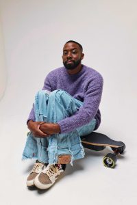 John Imah photographed for Galore in a purple sweater seated beside a skateboard