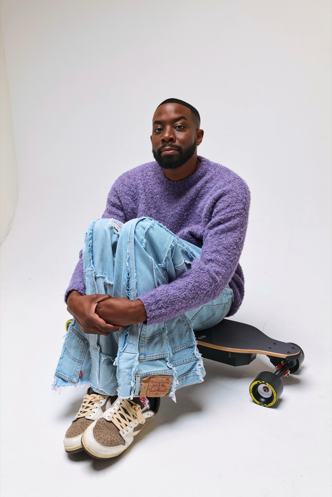 John Imah seated beside a skateboard in a studio fashion portrait